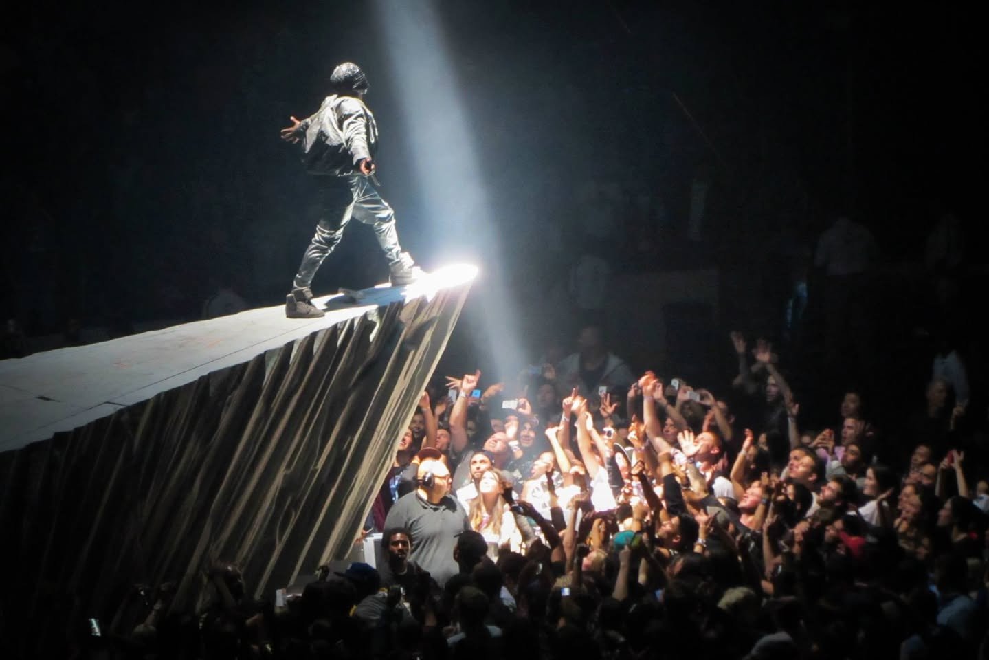 Kanye West: Yeezus Tour A performer on a stage wing under a spotlight above a large concert crowd with raised hands.