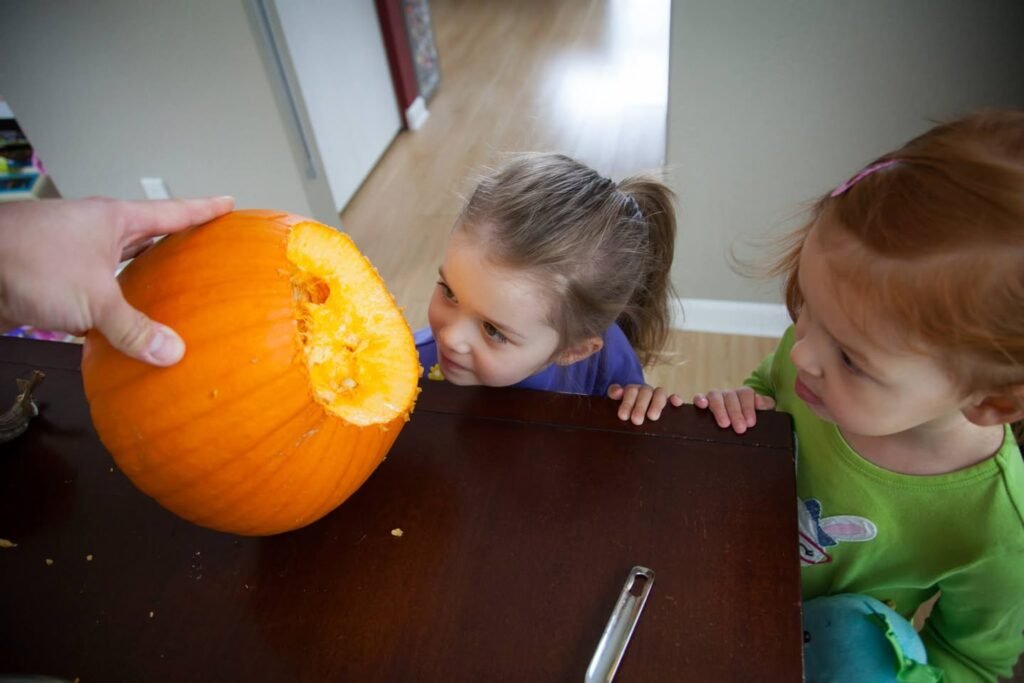 Two young children watch a person hold an orange pumpkin on a dark table.