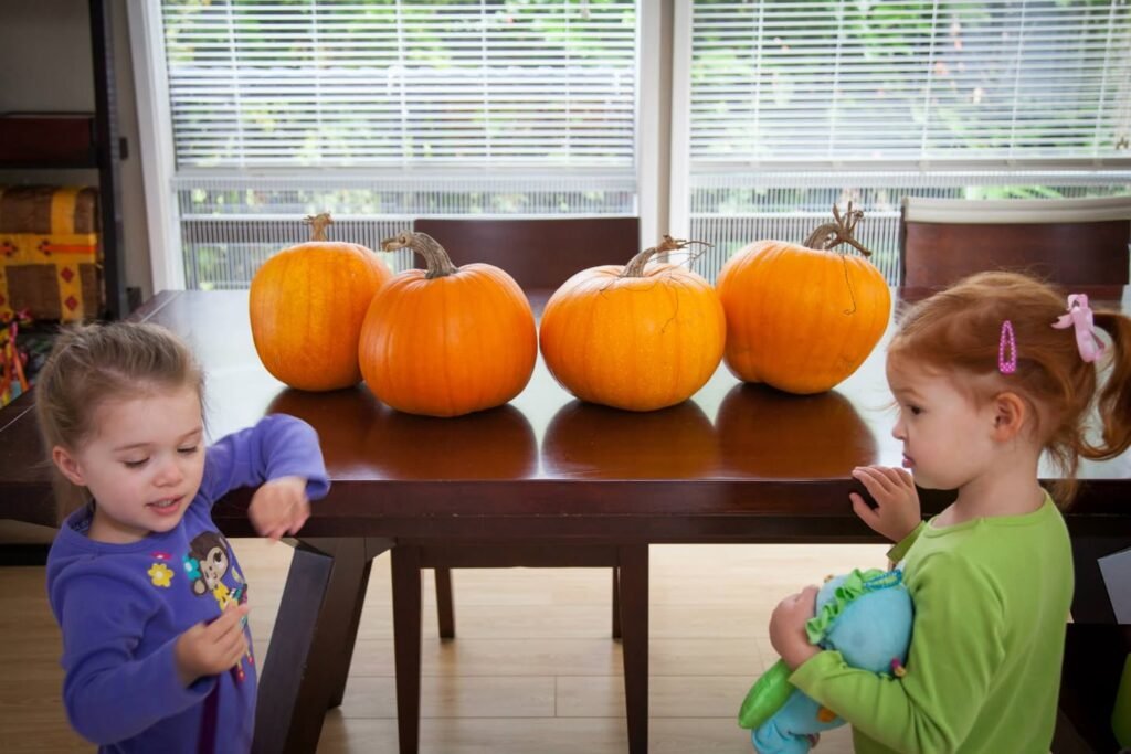 Two young children standing near a table with four bright orange pumpkins.