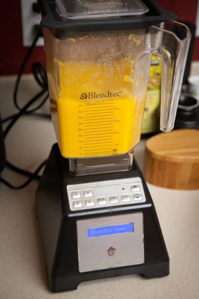 A Blendtec blender filled with a vibrant yellow smoothie sits on a kitchen counter next to a wooden container.