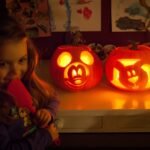 A child smiles beside glowing carved pumpkins in a cozy, dimly lit room.