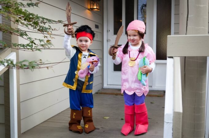 Two children dressed in matching Pinocchio and dinosaur costumes holding candy on a porch.