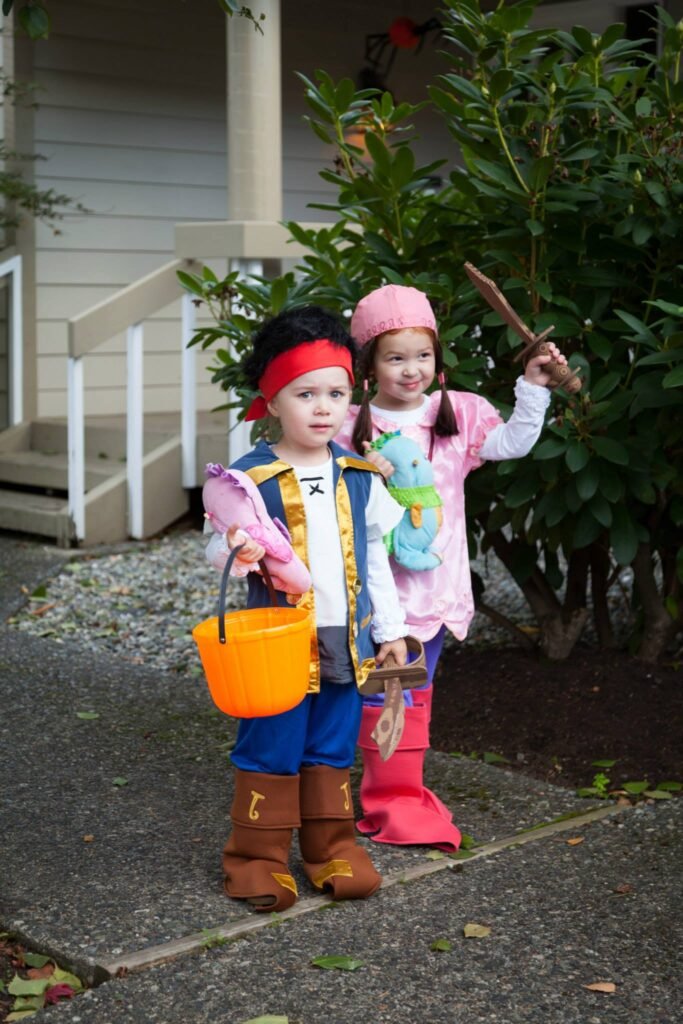 Two children in pirate and bunny costumes holding a bucket on a doorstep. One wears a red headband and blue jeans in brown boots.