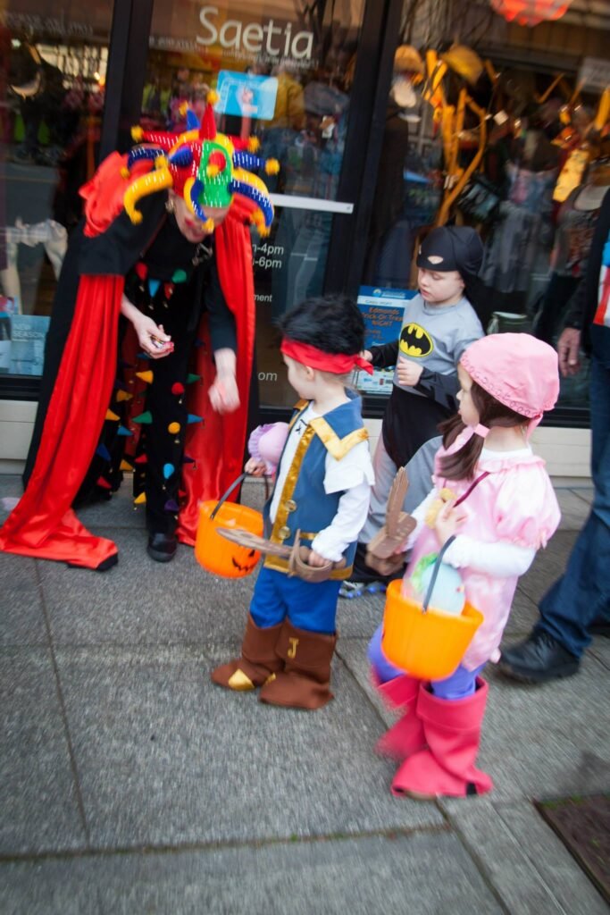 A group of costumed children holding orange tins talk to a tall clown figure outside a storefront. The kids are dressed as cartoon characters and stand in a neat line on a city sidewalk.