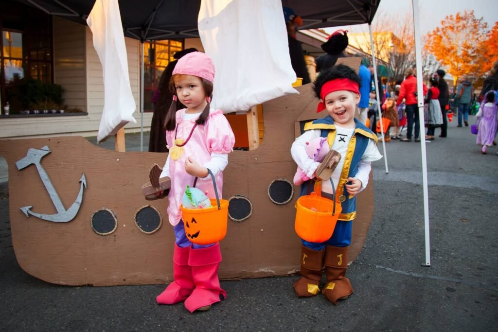 Two children dressed as pirates standing in front of a cardboard pirate ship display. They are holding plastic buckets for candy at an outdoor Halloween event.
