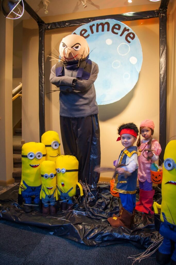 A young child dressed in a superhero costume stands proudly between life-size Minion characters and a towering Dr. Evil statue in a themed photo booth, captured back-to-back with a close-up of the characters.