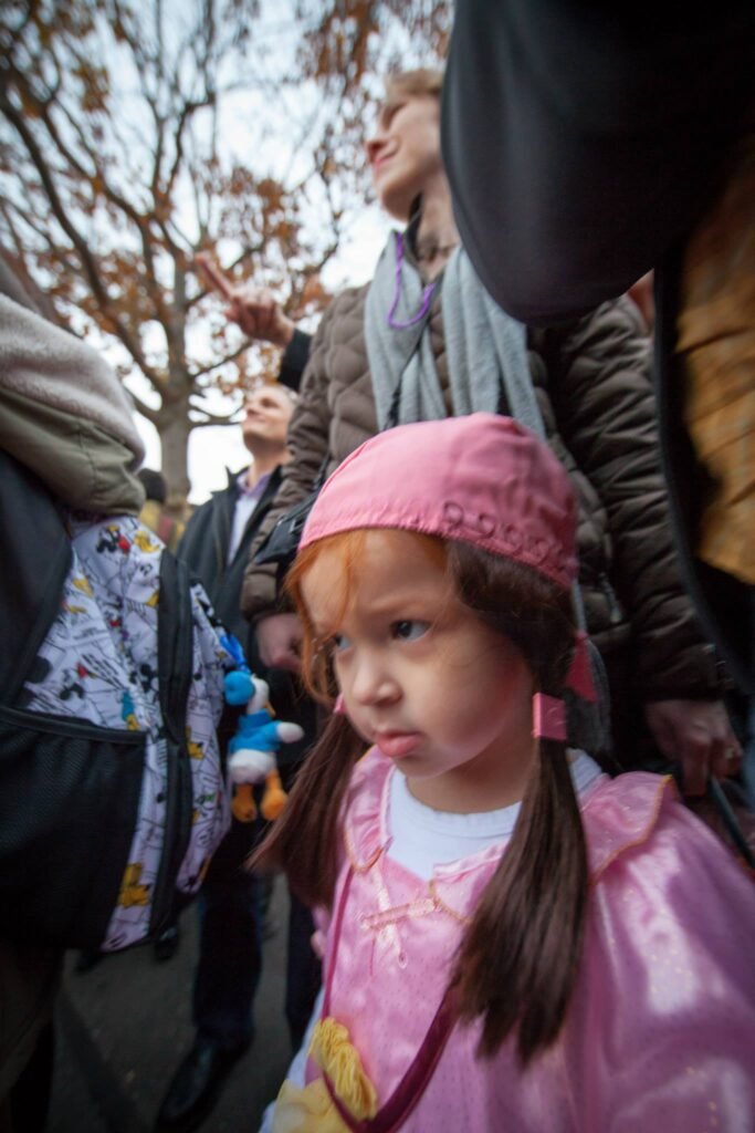 A young child in a pink hat and raincoat stands among a crowd of people outdoors on a cloudy day.