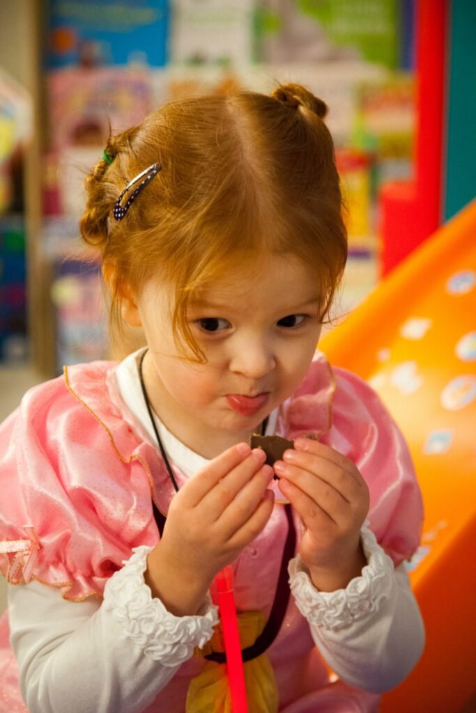 A young child in a pink costume looks closely at a small item. The background is blurred in a lively indoor environment.