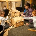 Children participate in a woodworking activity, constructing small wooden structures with an instructor's guidance. Close-up view of the wooden building materials.