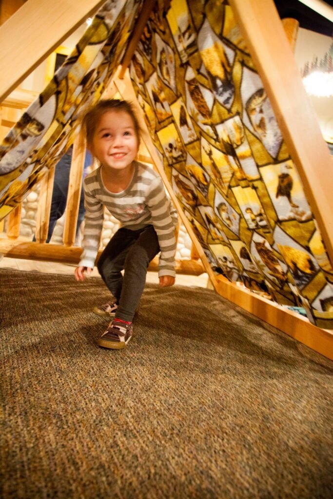 A child crouches inside a large triangular mosaic installation featuring diverse photographic tiles. The structure is situated on a textured carpet floor.