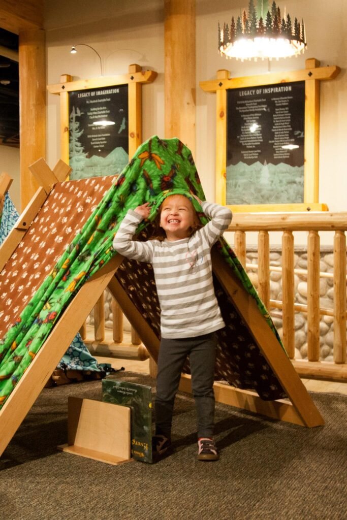A child explores a colorful tpee tent in a rustic lodge play area. Wooden beams and warm lighting add to the cozy atmosphere.