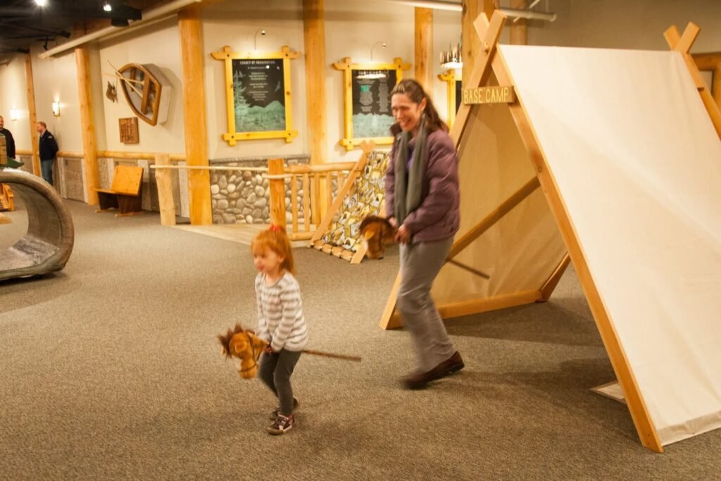 A toddler plays with a toy sword inside a children's museum exhibit. An adult accompanies the child in the interactive display area.