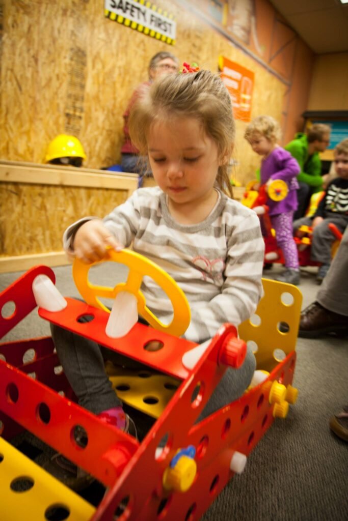 A child sits in a colorful construction ride-on toy with a yellow steering wheel. The background shows a room with a 'SAFETY FIRST' sign and a yellow hard hat.