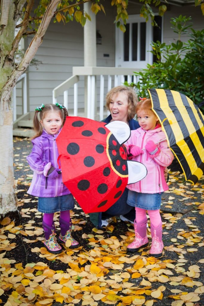 A family stands in a yard covered with autumn leaves, a small child in a pink raincoat holds a ladybug umbrella and another holds a black and yellow striped umbrella.