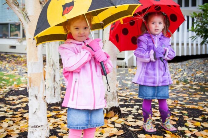 Two children hold bee and ladybug shaped umbrellas while standing on a ground covered in yellow autumn leaves.