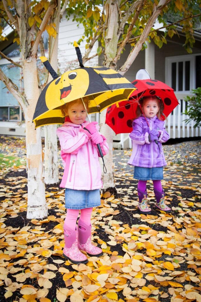 Two children hold bee and ladybug shaped umbrellas while standing on a ground covered in yellow autumn leaves.