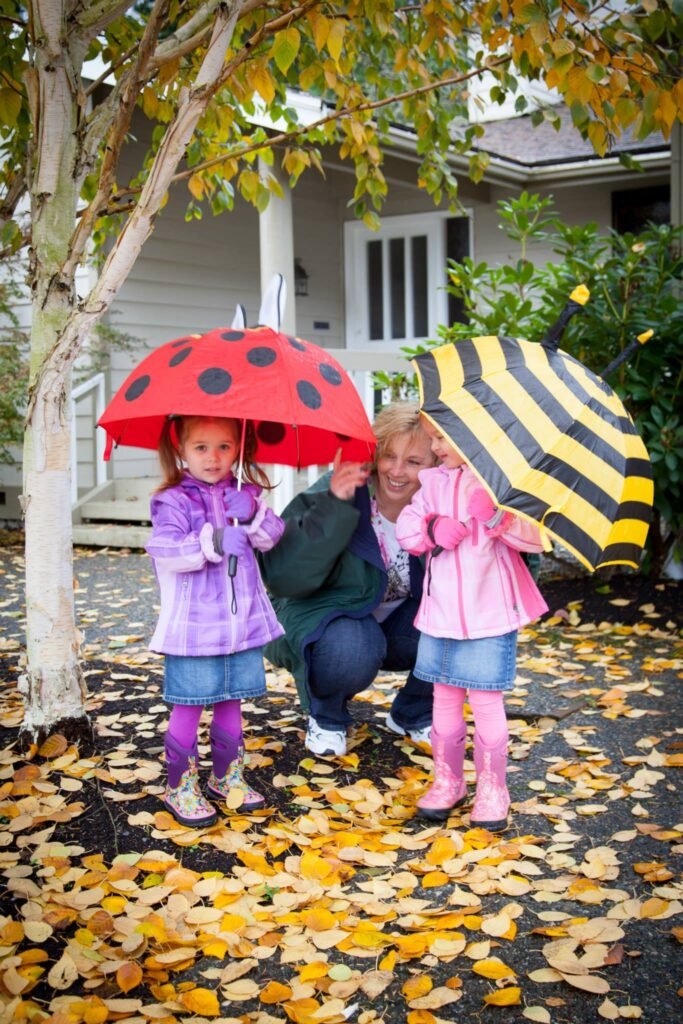 Two children holding colorful umbrellas, standing on a path covered with yellow leaves. A woman kneels beside them in front of a picturesque house.