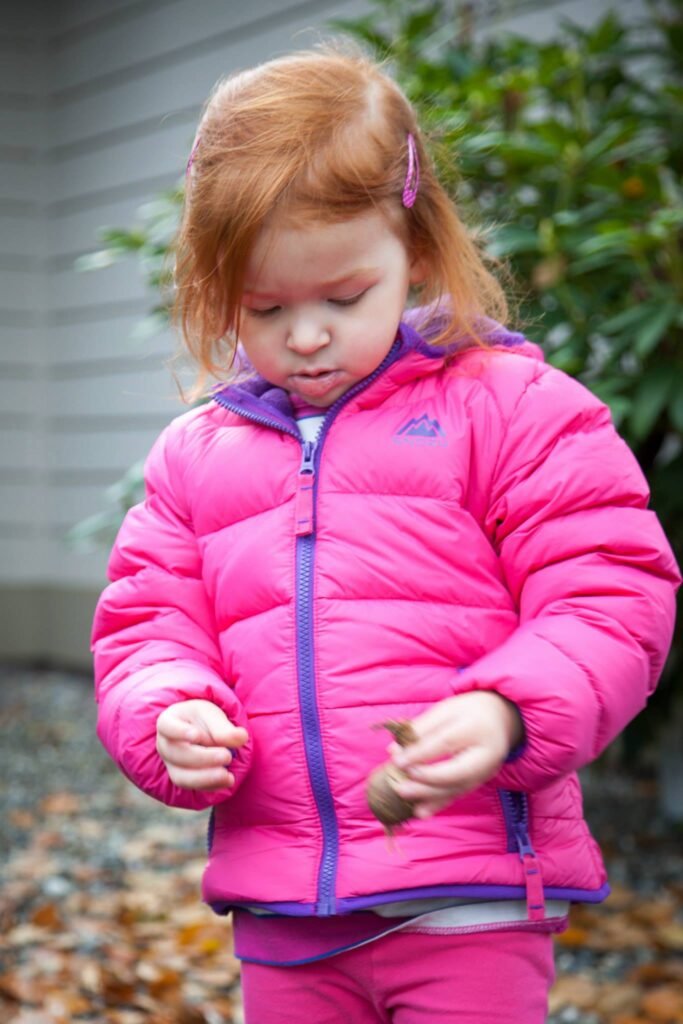 A child in a vibrant pink puffer jacket examines leaves on the ground outdoors.