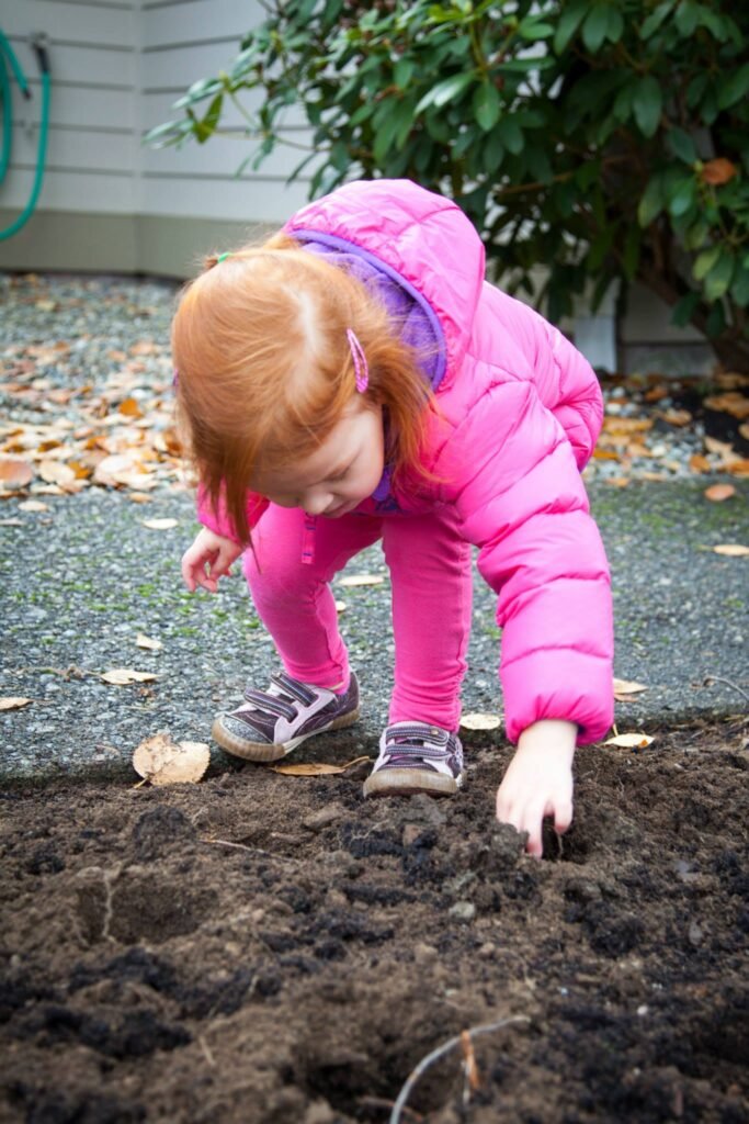 Child in pink clothing exploring fresh dirt outdoors. Curious hands reaching into soil near a garden bed.