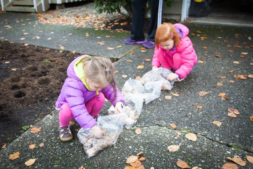 Two children in purple and pink winter jackets crouch on a stone patio, playing with plastic bags.