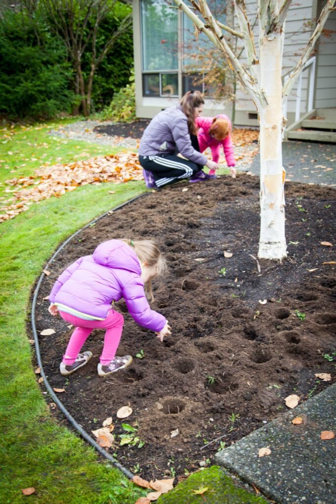 Children forming small zodiac balls in a garden bed near a tree. The scene is set in a Pacific Northwest outdoor space.