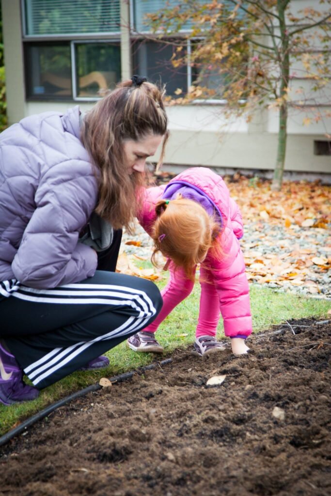 An adult and child in jackets tending to a garden bed with fallen leaves in the background. A young person in a pink coat and adult in a grey jacket are working in the soil.