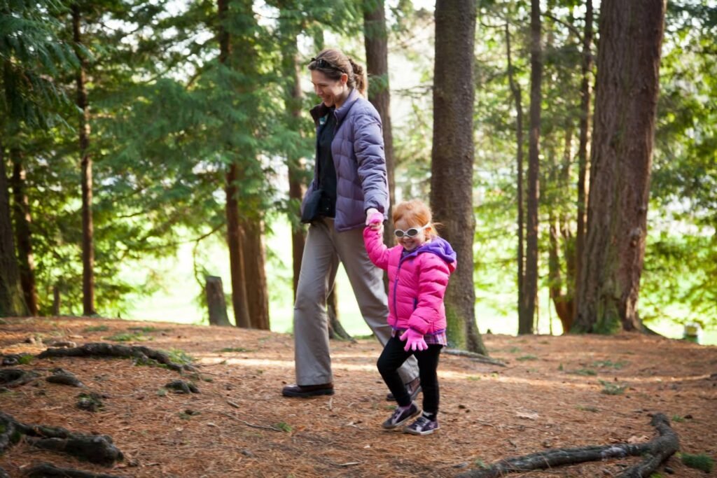 A person and a child walking through a forest. The forest features tall trees, a dirt floor, and a visible root system.