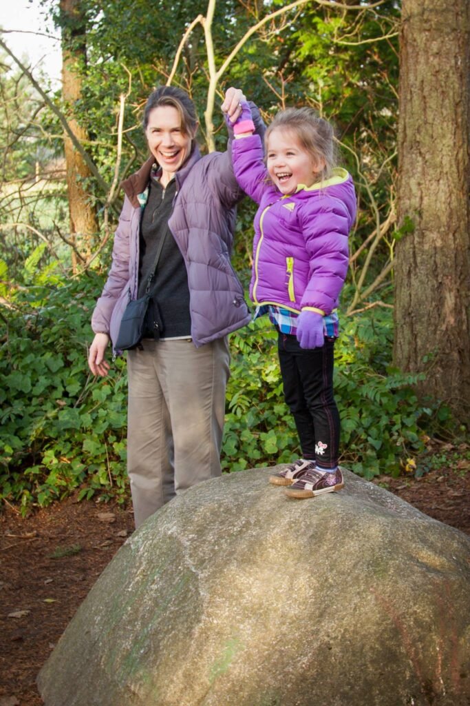 A woman and a young child in purple jackets stand together on a large boulder in a wooded area, sharing a happy moment.