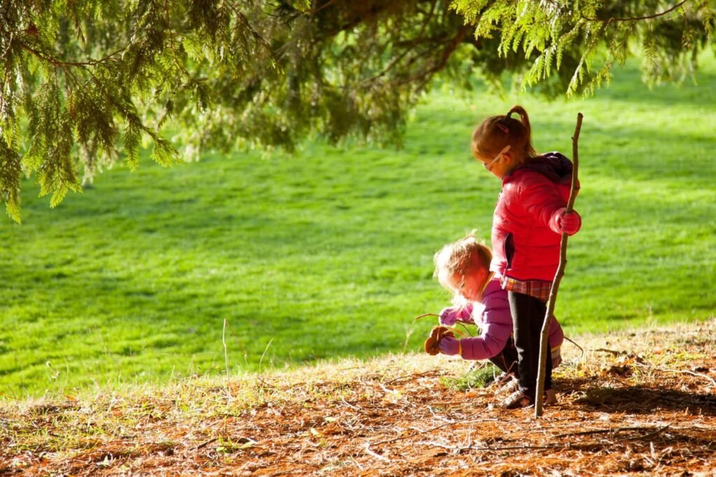 Two children explore a grassy park under a large tree with autumn leaves, holding a stick and crouching on the ground.