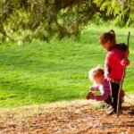 Two children explore a grassy park under a large tree with autumn leaves, holding a stick and crouching on the ground.