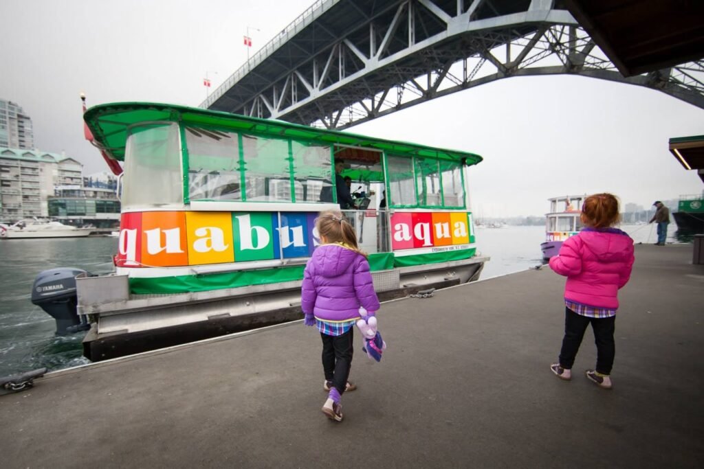 Children in colorful jackets walk along a pier beside a vibrant green and multi-colored water taxi with a large steel bridge in the background.