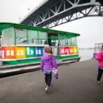 Children in colorful jackets walk along a pier beside a vibrant green and multi-colored water taxi with a large steel bridge in the background.