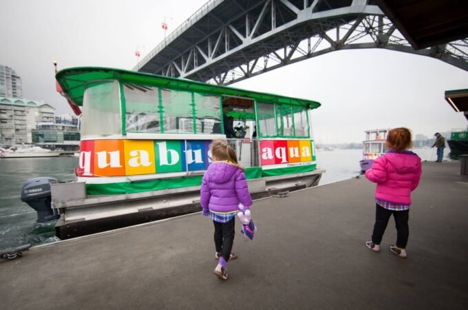 Children in colorful jackets walk along a pier beside a vibrant green and multi-colored water taxi with a large steel bridge in the background.