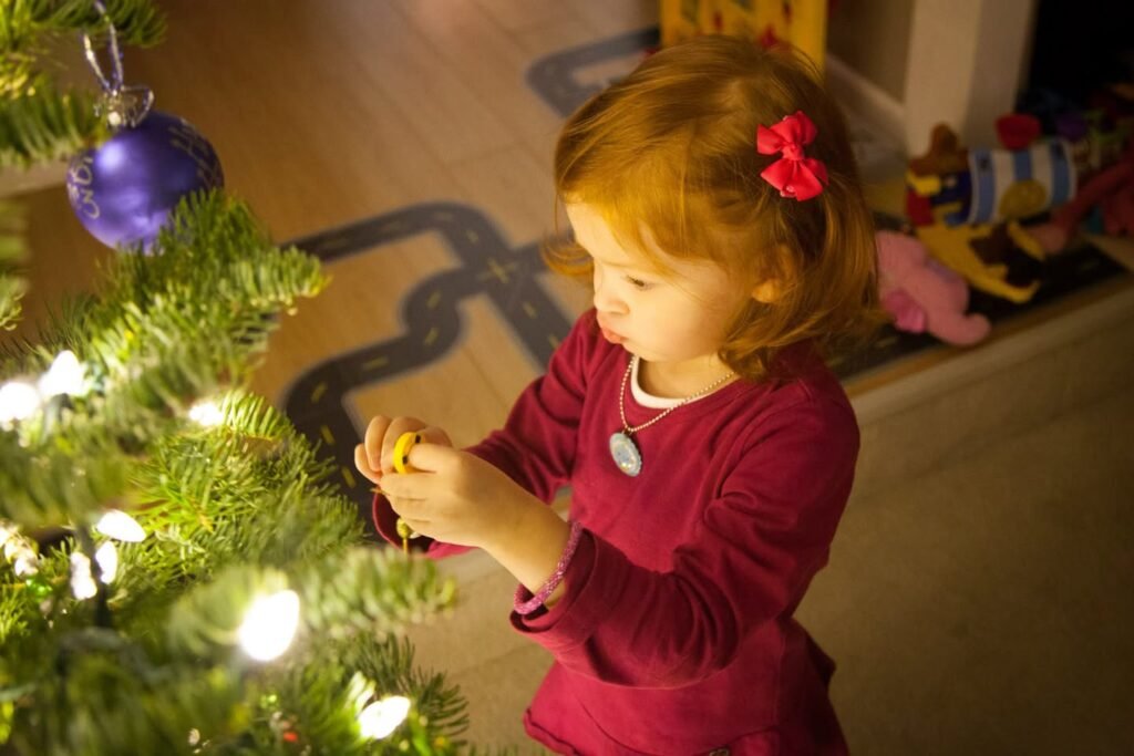 A young child in a red shirt hangs an ornament on a lit Christmas tree.
