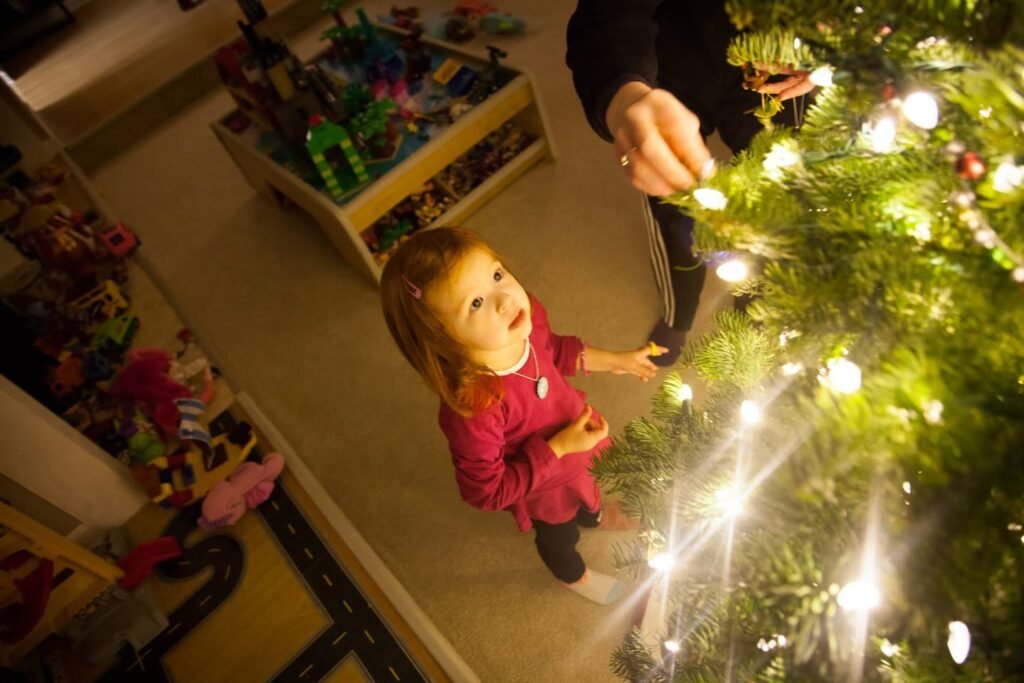 A child in a red top admiring a decorated Christmas tree with glowing lights, with a toy box in the background.