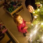 A child in a red top admiring a decorated Christmas tree with glowing lights, with a toy box in the background.