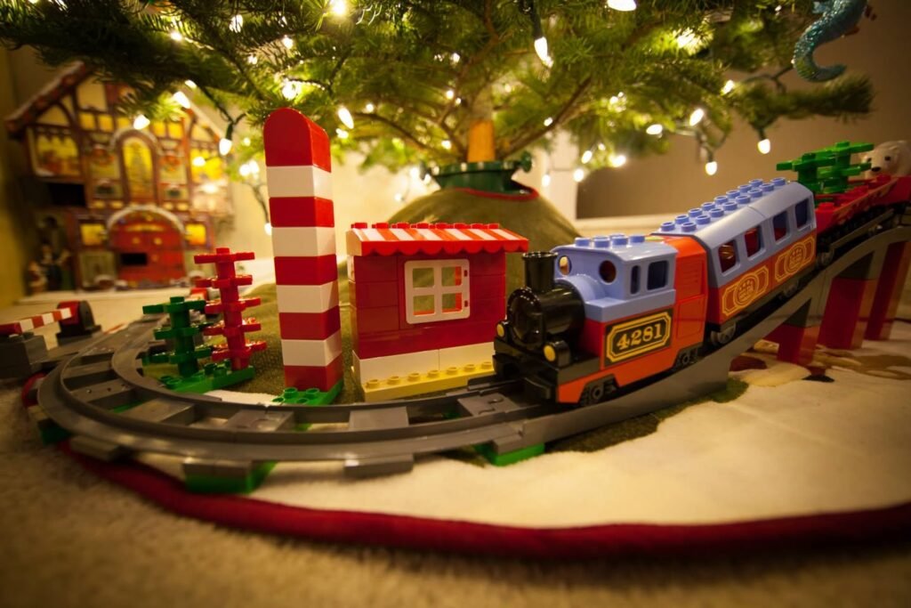 A colorful toy train set with red and green elements arranged under a Christmas tree with lit string lights.