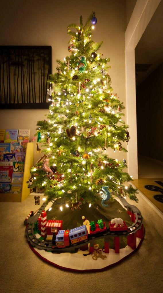 A warmly lit Christmas tree decorated with ornaments and lights, featuring a festive toy train set as its tree skirt.