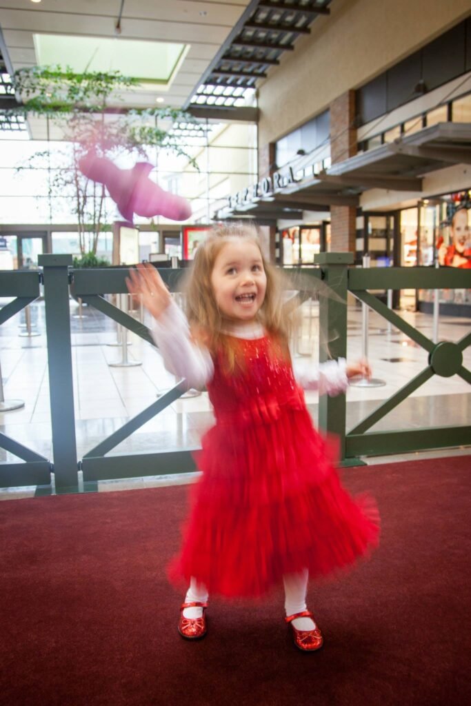 A child in a red dress is skipping joyfully on a red carpet inside a modern shopping mall.