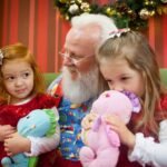 A cheerful visit with Santa Claus. Two young girls hold plush toys while Santa smiles warmly in a festive setting.