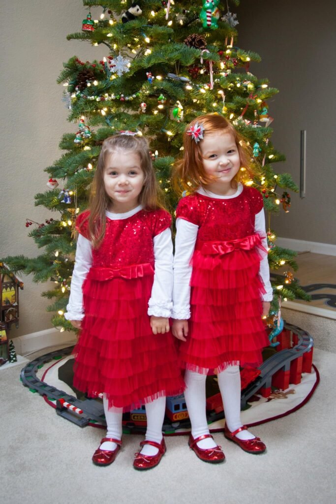 A pair of children stand in matching red ruffled dresses in front of a decorated Christmas tree. Below, several details of the festive scene are shown.