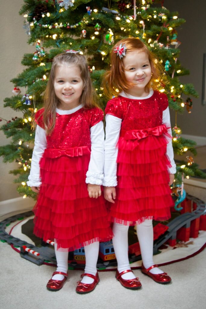Two sisters wearing matching red tiered dresses and white tights stand before a decorated Christmas tree. Close-up details show individual smiles and festive red shoes.