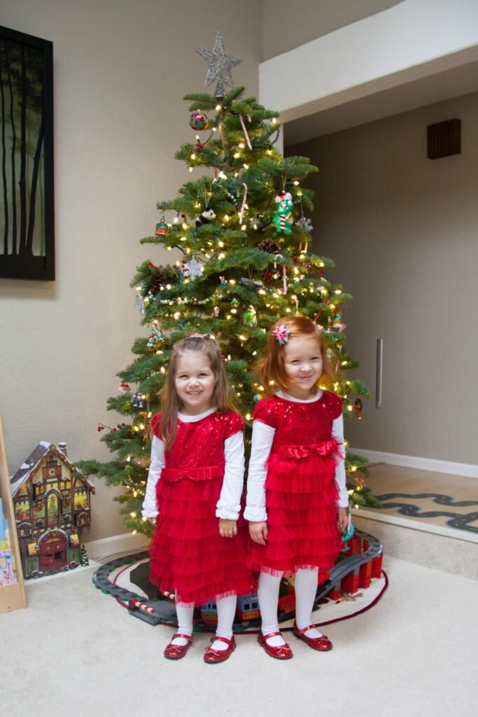 Children posing in red dresses next to a decorated Christmas tree in a home.