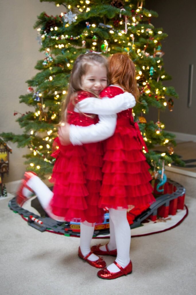 A young person in a red coat and white tights hugging another person in front of a lit Christmas tree with ornaments and a decorative tree skirt.