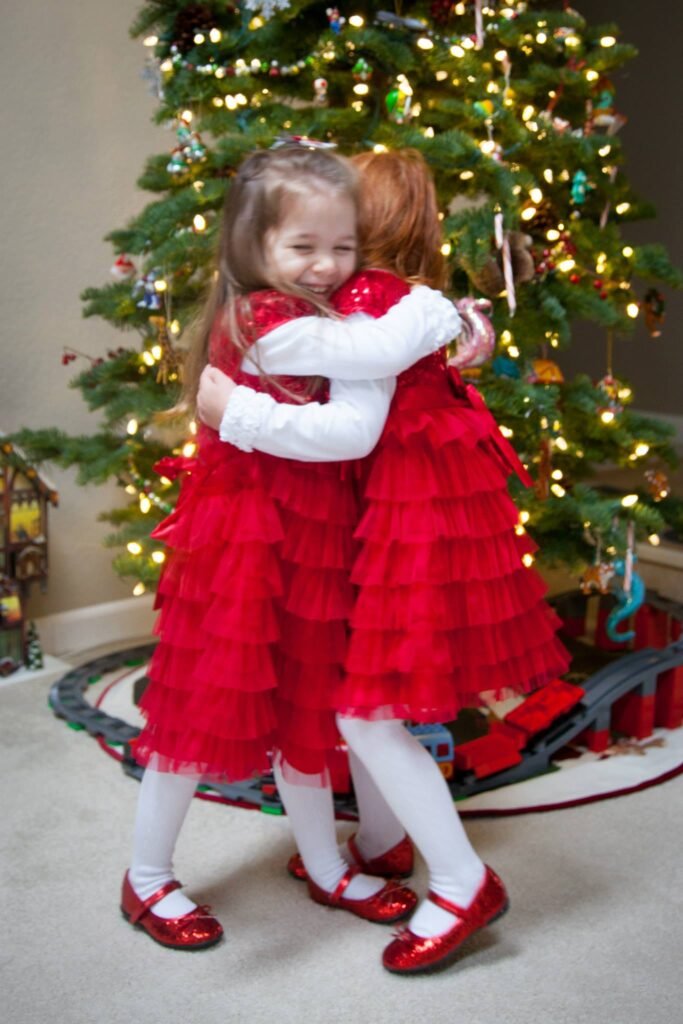 Two young girls in matching red tulle dresses and white tights share a warm hug in front of a decorated Christmas tree with glowing string lights.