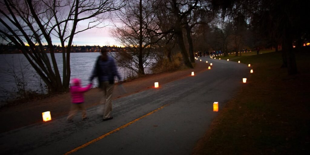 A paved path lined with glowing candles curls beside a calm lake at dusk. A person walks with a small child carrying a glowing candle near the shoreline.