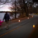 A paved path lined with glowing candles curls beside a calm lake at dusk. A person walks with a small child carrying a glowing candle near the shoreline.