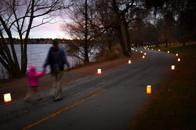 A paved path lined with glowing candles curls beside a calm lake at dusk. A person walks with a small child carrying a glowing candle near the shoreline.