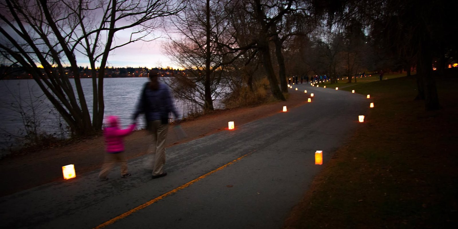A paved path lined with glowing candles curls beside a calm lake at dusk. A person walks with a small child carrying a glowing candle near the shoreline.