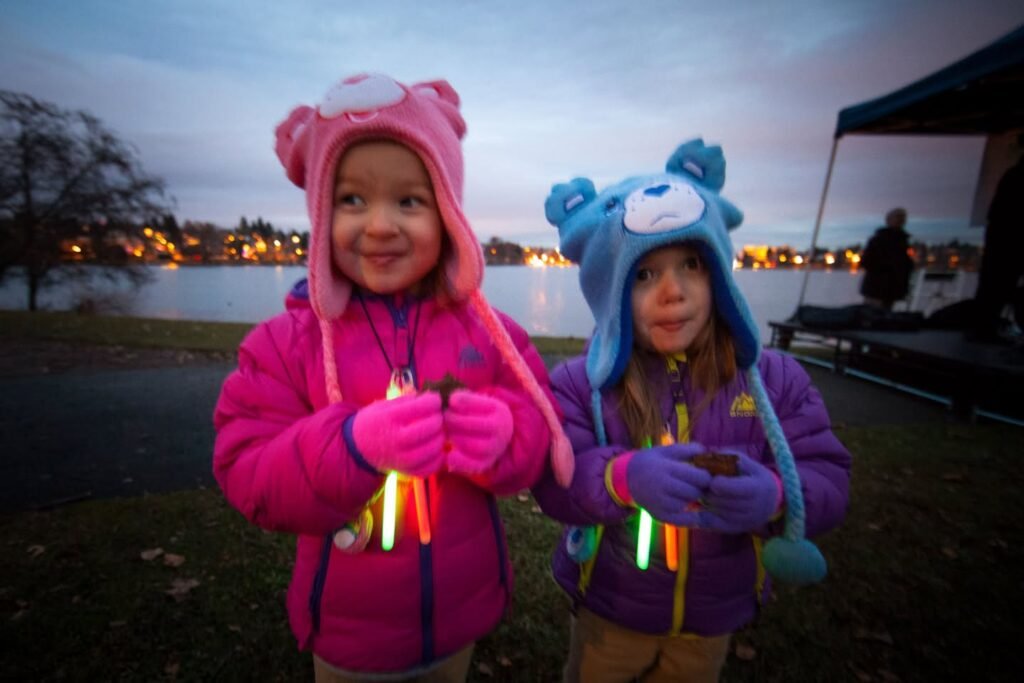Two individuals in colorful hooded outfits hold glowing sticks in front of a lake at twilight, with distant town lights visible.